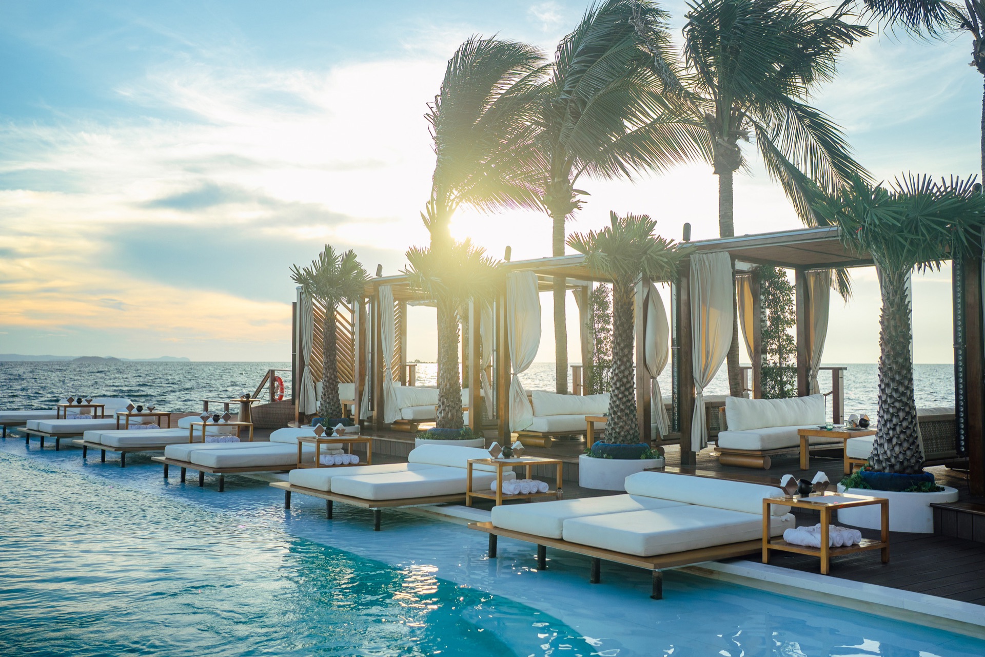 Golden hour — the sun setting through the palms over the glowing infinity pool